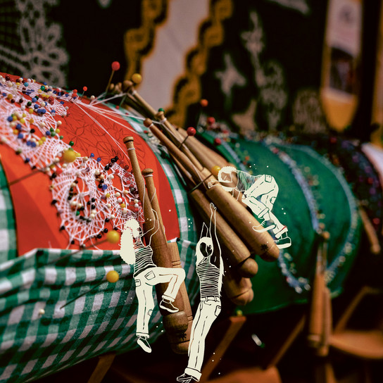 A display of colorful lace-making pillows with wooden bobbins, showcasing intricate lace designs. In the foreground, an overlay illustration of two children playfully climbing on the lace structure.