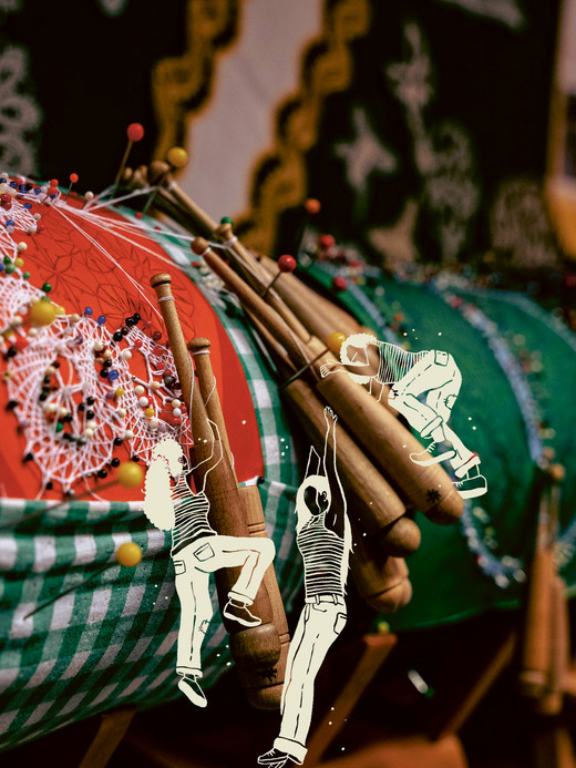 A display of colorful lace-making pillows with wooden bobbins, showcasing intricate lace designs. In the foreground, an overlay illustration of two children playfully climbing on the lace structure.