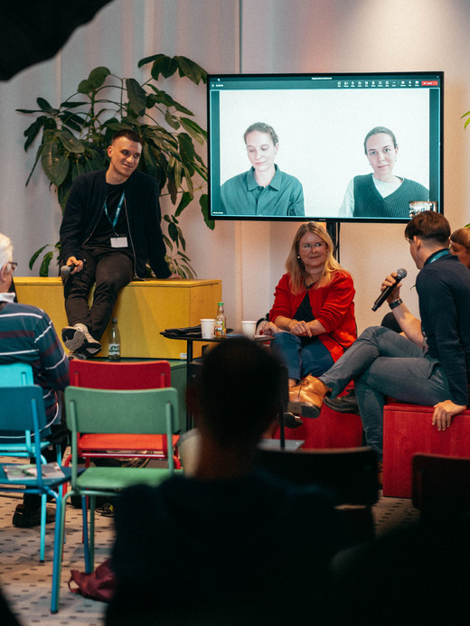 A group of people sit and engage in a discussion in a room with colorful chairs and potted plants. A screen in the background displays a video call with two individuals, and another screen shows text. Several attendees face the discussion panel.