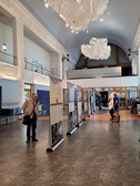 A man reads an exhibition board in a spacious hall with high ceilings, modern lighting and hanging fabric decorations. Other people can be seen in the background, some standing, others sitting.