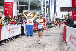 A male runner crosses the finish line of a race, cheered on by a crowd and a person in a red and white mascot costume. A digital clock shows the race time of 2:54:55. Event banners line the route.