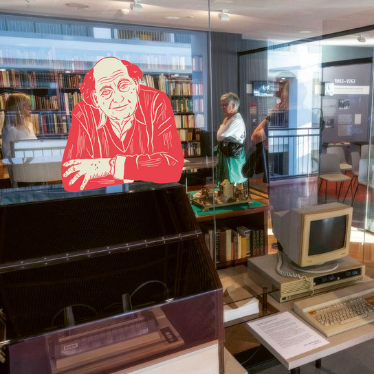 A museum exhibit featuring vintage computer equipment and a digital artwork of an older man in a red outline. Visitors are observing the displays, and shelves filled with books are visible in the background.