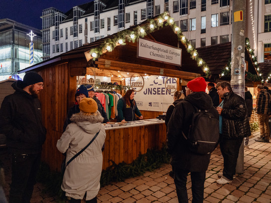 People gather around a festive wooden stall decorated with fairy lights at a night market in Chemnitz. The sign at the top reads "Chemnitz European Capital of Culture 2025". Some browse through the offerings while others chat nearby. Buildings can be seen in the background.