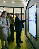 Ein Mann im Anzug demonstriert zwei für kaltes Wetter gekleideten Frauen ein Touchscreen-Display. Sie befinden sich in einem Gebäude, im Hintergrund sind Banner und Schilder zu sehen, die auf kulturelle Veranstaltungen hinweisen. Das Display bietet mehrere Optionen und die Gruppe scheint interessiert.