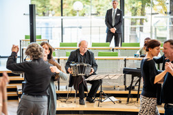 Ein Musiker spielt auf der Bühne ein Bandoneon, während mehrere Paare um ihn herum tanzen. Ein Mann im Anzug steht im Hintergrund und beobachtet die Szene. Der Schauplatz scheint eine moderne Halle mit großen Fenstern zu sein.
