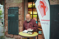 An older man with glasses and a beard stands at a round table in front of a building and reads an open book, next to which other books are piled up. Nearby are chalkboards and a white flag with red lettering and a logo.