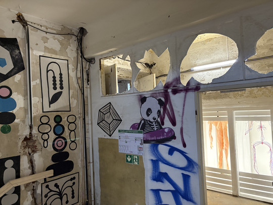 A room with peeling paint, geometric wall design and graffiti with a panda on a purple bumper car. There are exposed wires, emergency exit signs and a window with cut-out arches above the door.
