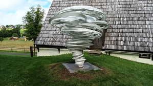 Eine Metallskulptur in Form eines wirbelnden Tornados steht auf einem grasbewachsenen Rasen vor einem Holzgebäude mit Schindeldach. Im Hintergrund sind Bäume und ein Zaun zu sehen.