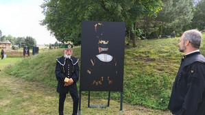 Zwei Männer in historischen Kostümen stehen im Freien neben einer schwarzen Schautafel mit dem stilisierten Bild einer Person, die eine Krone und verzierte Kleidung trägt. Im Hintergrund sind Bäume und Gras zu sehen.