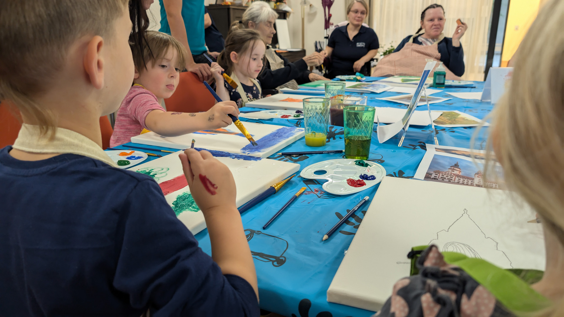 Eine Gruppe von Kindern und Erwachsenen sitzt um einen Tisch herum und malt. Der Tisch ist mit blauem Plastik bedeckt und verschiedene Malutensilien, darunter Farbpaletten und Pinsel, sind darauf verstreut.