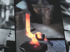 Eine Nahaufnahme eines glühenden, erhitzten Metallstücks, das mit einer Zange auf einem Amboss geschmiedet wird. Sie zeigt die orange-rote Farbe des Metalls, während es in einem Schmiedeprozess geformt wird.