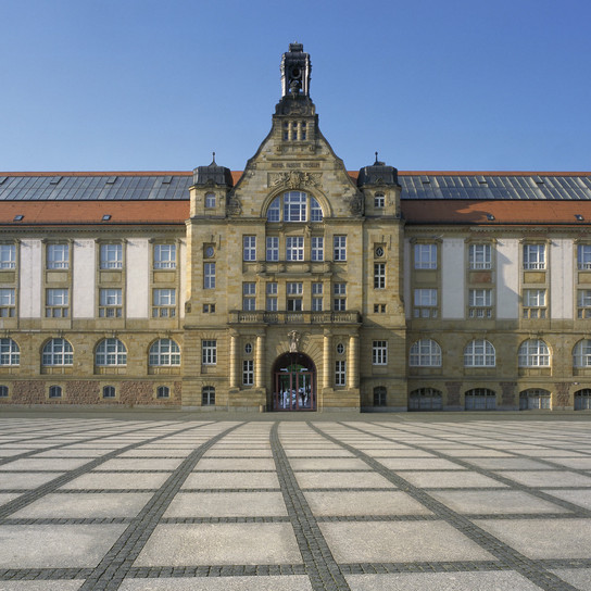 A large historic building with an ornate facade, featuring arched windows, a central clock tower, and a red-tiled roof, stands against a clear blue sky. The foreground consists of an expansive, checkerboard-patterned plaza made of stone tiles.