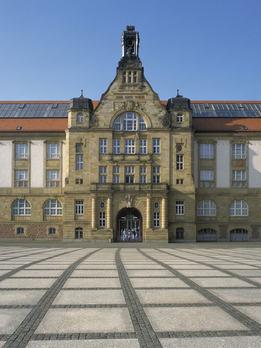 A large historic building with an ornate facade, featuring arched windows, a central clock tower, and a red-tiled roof, stands against a clear blue sky. The foreground consists of an expansive, checkerboard-patterned plaza made of stone tiles.