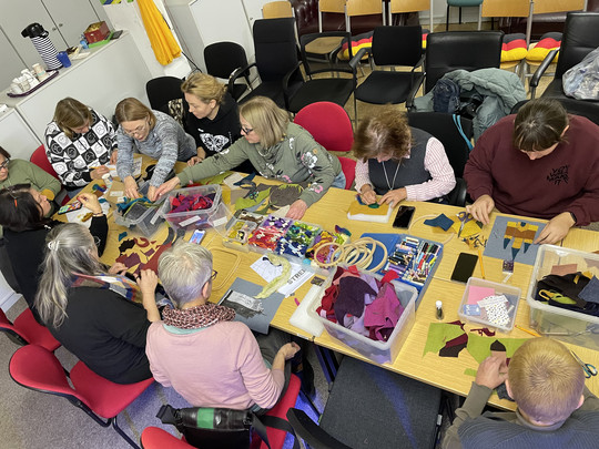 Eine Gruppe von Menschen sitzt um einen Tisch und beschäftigt sich mit Kunsthandwerk. Verschiedene Materialien wie Stoff, Scheren und Klebstoff liegen verstreut auf dem Tisch. Der Raum ist mit Stühlen, Tassen und farbenfrohen Dekorationen ausgestattet. Jeder wirkt konzentriert und in seine Arbeit vertieft.