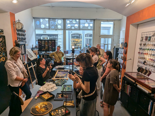 A group of people are gathered in an art shop. Some are standing, while others are seated around a table filled with craft materials. Shelves filled with art supplies line the walls. The atmosphere appears collaborative and focused.