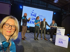 A woman takes a selfie at an indoor event. In the background, four people are standing on a stage in front of a large screen showing "The Unseen Chemnitz European Capital of Culture 2025". The setting looks festive and professional.