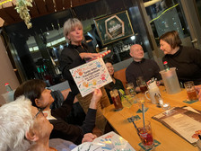Eine Gruppe von Menschen sitzt um einen Restauranttisch, lächelt und unterhält sich. Eine Frau hält ein geschmücktes Schild mit der Aufschrift "Danke liebes Freundeskreispubteam!" Getränke und Speisekarten stehen auf dem Tisch. Die Stimmung ist fröhlich und feierlich.