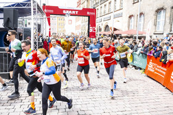 A group of runners in colourful outfits take part in a city run that begins under a red arch with the inscription "START", while spectators line the cobbled street in a historic district.
