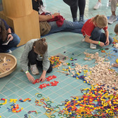 Children sit and kneel on a blue-tiled floor, sorting and arranging colourful geometric shapes and wooden pieces into patterns. Adults and other children stand nearby and watch or take part in the activity.