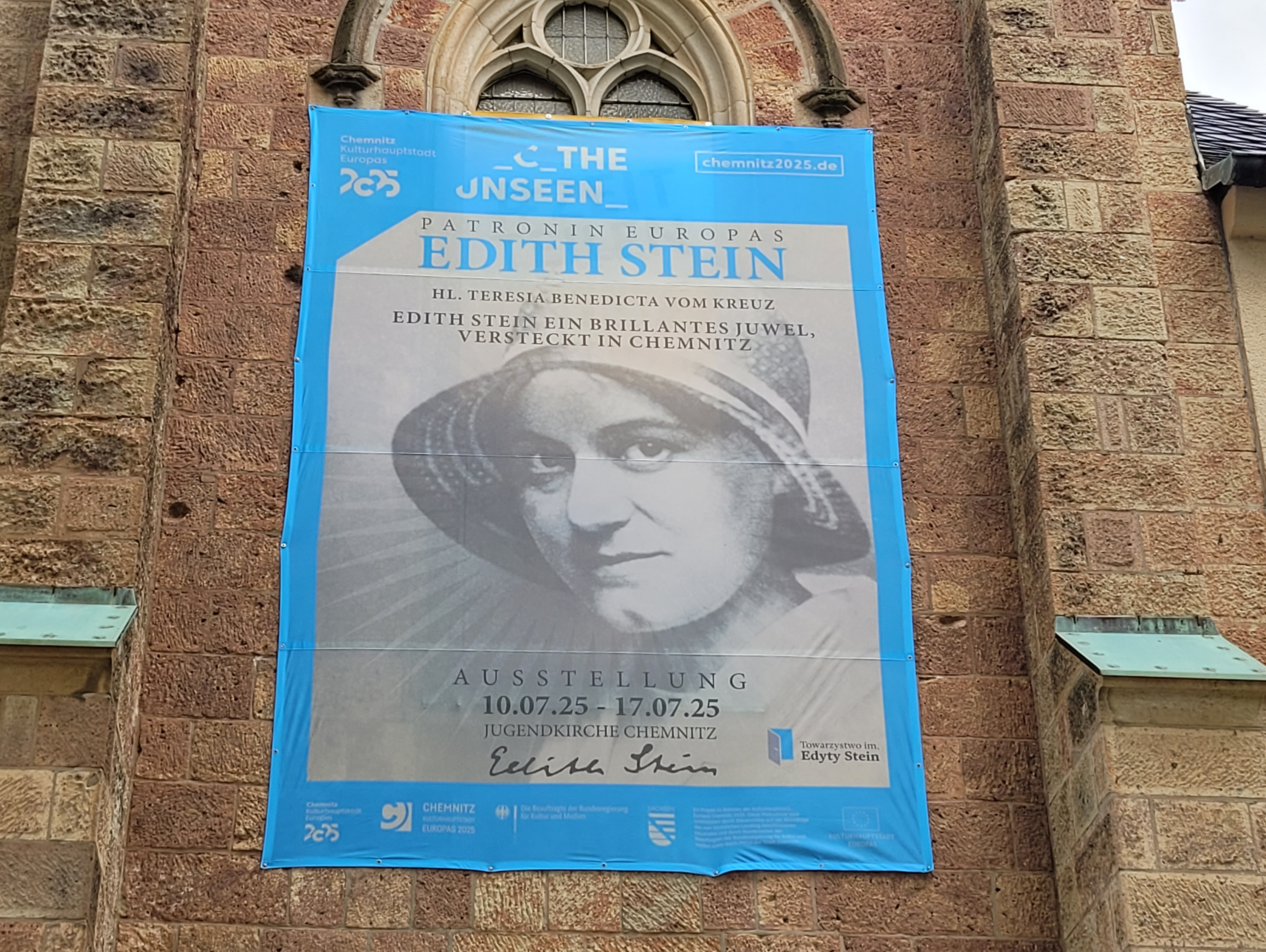 A black and white portrait of Edith Stein with a hat is depicted on a blue banner on a brick church wall. The text announces an exhibition about her entitled "The Invisible" in Chemnitz, Germany, from 10 July 2025 to 17 July 2025.