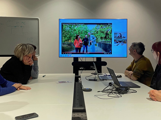 Sieben Personen sitzen an einem Konferenztisch und verfolgen eine Präsentation auf einem großen Bildschirm. Der Bildschirm zeigt ein Foto von einem Paar, das auf einer Brücke steht. Auf dem Tisch liegen ein Laptop und diverse Kabel. Im Hintergrund ist ein Whiteboard mit Notizen zu sehen.