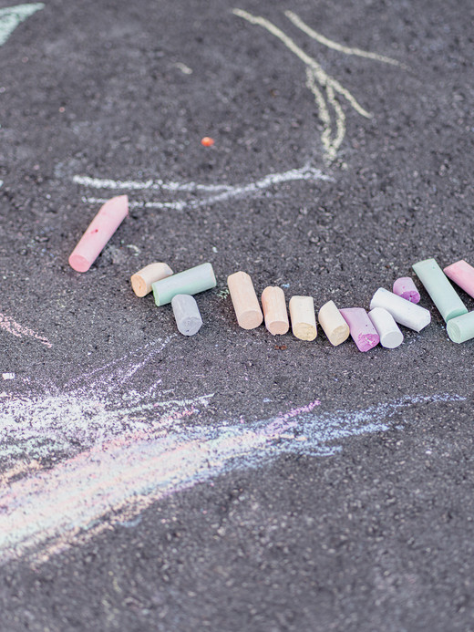 Bunte Kreidestücke liegen auf dem Asphalt verstreut, umgeben von Kreidezeichnungen, darunter ein Teil eines großen Cupcakes und abstrakte Formen.