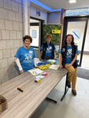 Three smiling volunteers in blue T-shirts stand behind a registration table with ID cards, pens and flyers in a foyer. A banner and glass doors can be seen in the background.