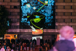 Eine Menschenmenge beobachtet eine nächtliche Projektion auf ein Gebäude, die bunte abstrakte Formen, Text und einen roten Stern zeigt. Die Szene ist lebhaft, mit versammelten Menschen und einigen verschwommenen Figuren im Vordergrund.