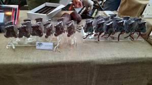 Schokoladenbären in Form von Lutschern werden an einem Stand auf einem Markt mit einer Tischdecke aus Sackleinen ausgestellt. Im Hintergrund sind weitere Schokoladenleckereien und eine Person mit Handschuhen zu sehen.