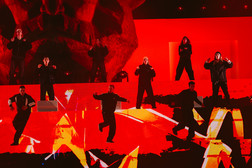 Nine performers dressed in black dance energetically on a stage with a bright red and orange backdrop, abstract patterns and dramatic lighting.