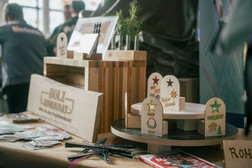 Ein hölzerner Auslagetisch mit handgefertigten Holzschildern, sternförmigen Ornamenten, Broschüren und kleinen dekorativen Tafeln mit deutschem Text, aufgestellt auf einem Markt oder einer Messe mit Menschen im Hintergrund.