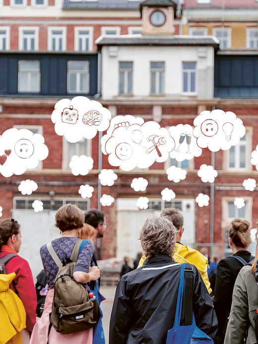 Eine Gruppe von Menschen steht im Freien vor einem historischen Gebäude. Über ihnen sind Sprechblasen mit verschiedenen Symbolen wie Smileys, einem Gehirn und einem Herz zu sehen, was darauf schließen lässt, dass sie über verschiedene Ideen oder Gefühle nachdenken.