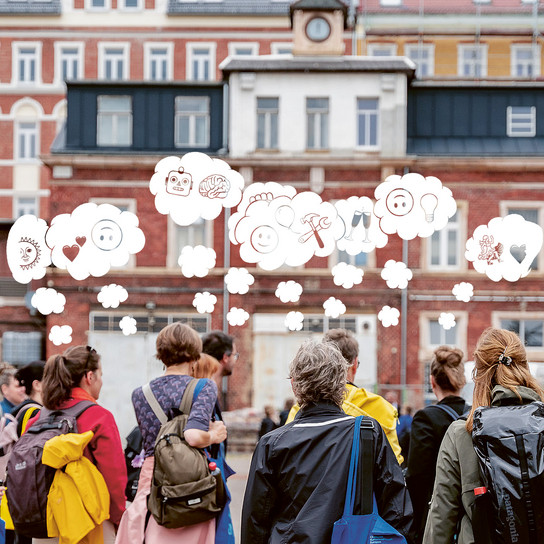 A group of people with backpacks stand facing a historic building. Thought bubbles above their heads contain various emojis, including a heart, brain, smiley, and musical notes, suggesting a diversity of thoughts and emotions.