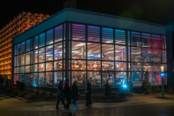 Ein modernes gläsernes Gebäude erstrahlt nachts in bunten Lichtern und ist innen mit hängenden Sternen geschmückt. Draußen auf dem Bürgersteig gehen mehrere Menschen vorbei. Die Atmosphäre wirkt lebendig und festlich.