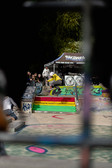 A skateboarder in a yellow hoodie performs a trick on a painted staircase in a skate park, watched by spectators. The ramps are decorated with graffiti and a tent with banners can be seen in the background.