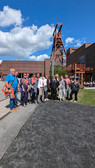 Eine Gruppe von Menschen steht an einem sonnigen Tag im Freien und posiert für ein Foto. Sie stehen vor einem Industriegebäude mit hohen, rotbraunen Gebäuden im Hintergrund. Der Himmel ist klar mit ein paar weißen Wolken. Einige Menschen im Hintergrund gehen spazieren und entspannen sich.