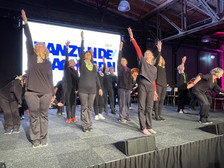 A group of older adults dressed in black perform a synchronised dance on stage, raising one arm in the air. A large illuminated sign and an industrial-style ceiling with visible beams can be seen in the background.