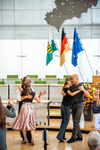 Zwei Paare tanzen eng beieinander in einem modernen Gebäude mit Fahnen (grün, deutsch, EU) im Hintergrund. Ein paar Leute schauen zu und machen Fotos; eine Frau in einem rosa Rock ist eine der Tänzerinnen.