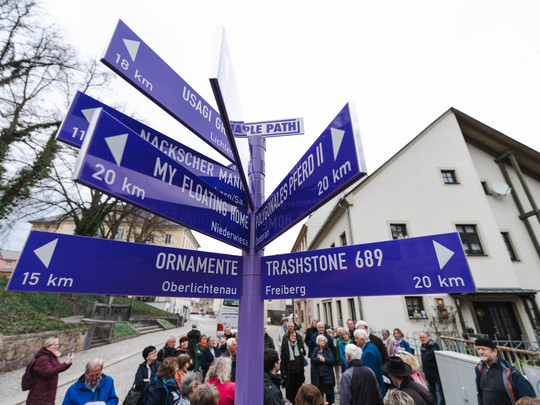 A group of people stand around a purple signpost at the PURPLE PATH in Hainichen with several arrows pointing in different directions, each labelled with destination names and distances in kilometres.