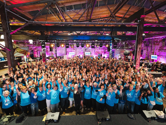 A large group of people in matching blue shirts stand together in a spacious, industrial-like room with exposed beams and colourful stage lights, smiling and waving at the camera.