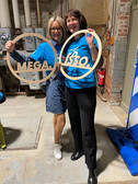 Two women are standing on a concrete floor in an interior room, smiling and holding round wooden signs with the words "MEGA" and "LABO". Both are wearing blue shirts; one is wearing a denim dress with glasses, the other black trousers. Behind them are exposed pipes and brick walls.
