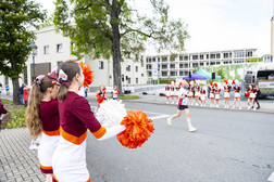 Cheerleaders in maroon and white uniforms with orange pompoms stand on a pavement and cheer on a runner in the street, while more cheerleaders gather in the background on the other side of the street.