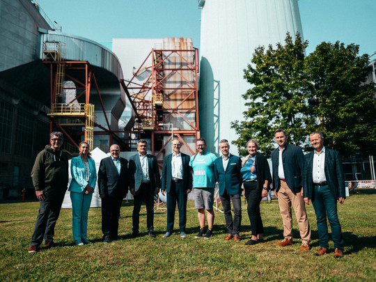 A group of ten people are standing outside on a lawn in front of an industrial backdrop. The group consists of men and women in business and leisure wear. They pose in a row and smile at the camera, with a sunny sky and trees in the background.