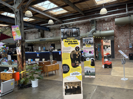 A covered event space with exposed brick walls and industrial beams features information boards, retro furniture, potted plants and people in the background. Sunlight shines through the skylights on the ceiling.