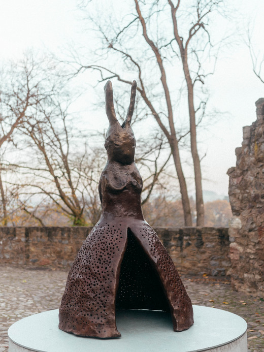 Eine Kaninchenstatue mit strukturierter Oberfläche steht auf einem runden Sockel im Freien. Das Kaninchen hat eine hohe, verzierte rockartige Struktur. Im Hintergrund sind kahle Bäume und eine Steinmauer zu sehen, was an einen Park oder eine historische Stätte erinnert.