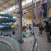 People gather in a modern indoor pool with high ceilings, natural light and a large, green water slide with a spiral. Some stand at empty pools, others sit at tables decorated with colourful banners.