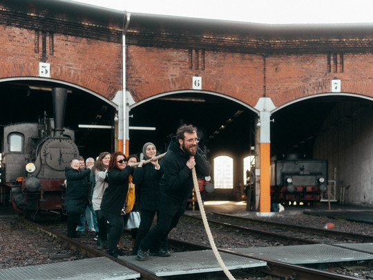 A group of people playfully pull an old steam locomotive out of an engine shed using a thick rope. The engine shed has several open compartments and the scene conveys a fun, communal effort.