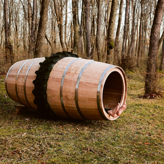 A large wooden barrel with metal bands is propped on its side in a forest clearing. An old tractor tire rests nearby. Surrounding the scene are leafless trees and brown grass, indicating late autumn or early winter.