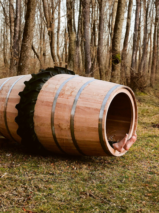 A large wooden barrel with metal bands is propped on its side in a forest clearing. An old tractor tire rests nearby. Surrounding the scene are leafless trees and brown grass, indicating late autumn or early winter.