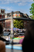 A crowd watches a skateboarding event in an outdoor skate park, with colourful ramps and banners visible. Historic red brick buildings and trees under a blue sky can be seen in the background.
